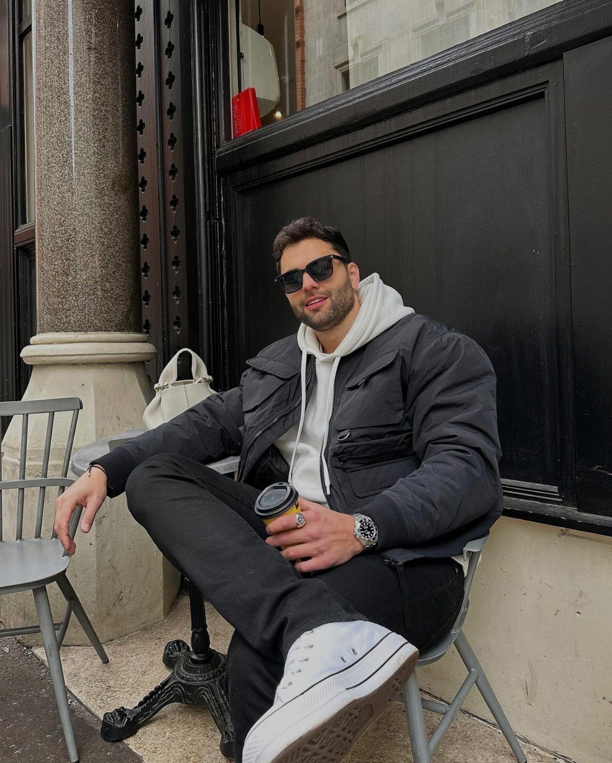 A man in a black jacket and white hoodie sitting at an outdoor cafe table.