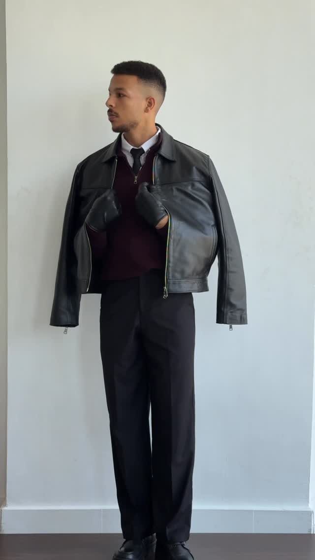 Man wearing a burgundy sweater, tie, black trousers, and a black leather jacket draped over his shoulders.