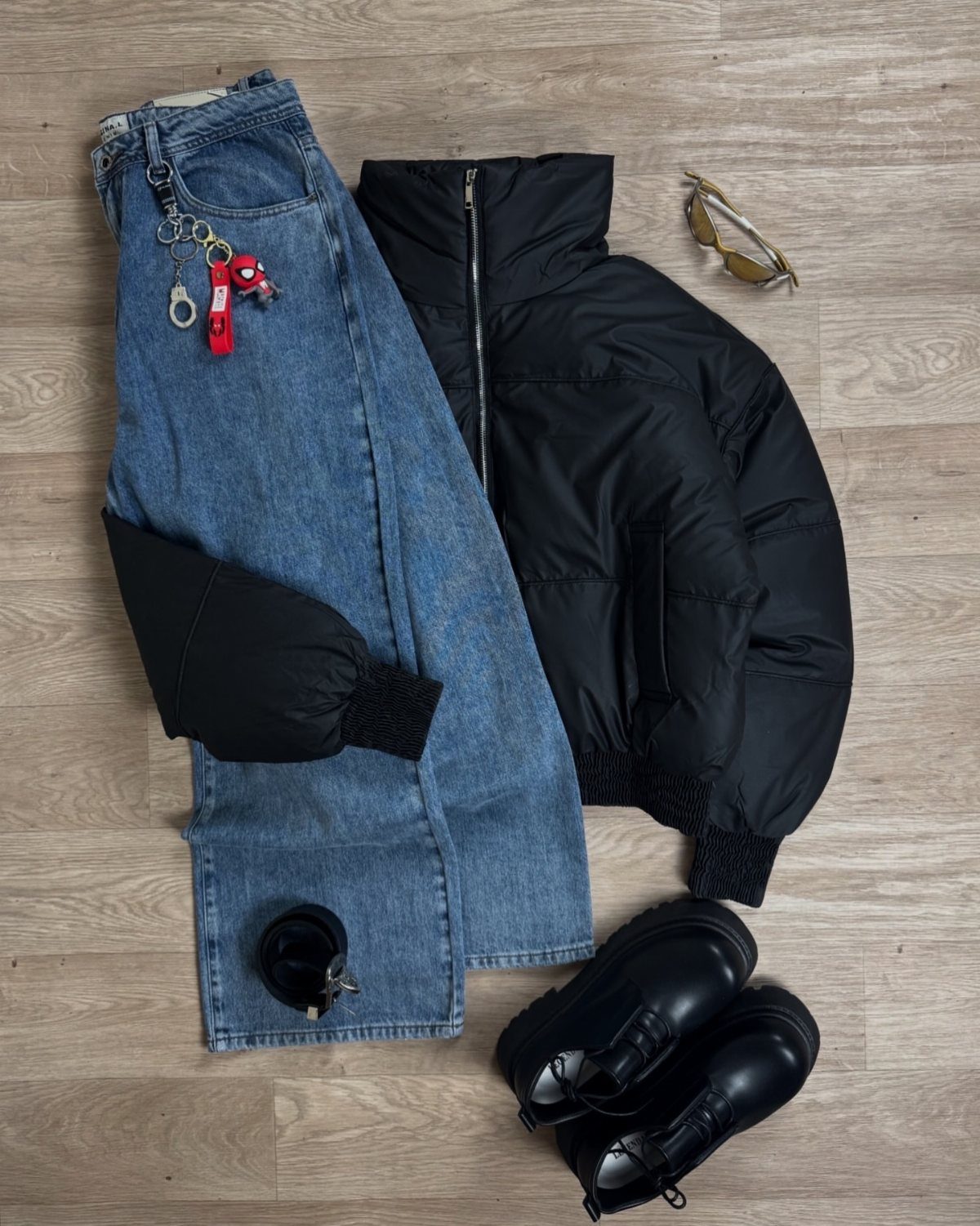 Black puffer jacket, blue jeans, black boots, and sunglasses arranged on a light wooden floor.