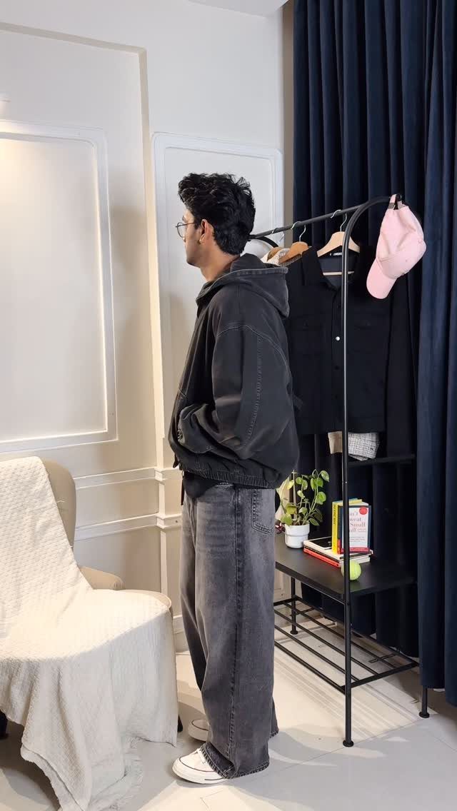 Man in a black hoodie and grey jeans standing near a clothing rack.