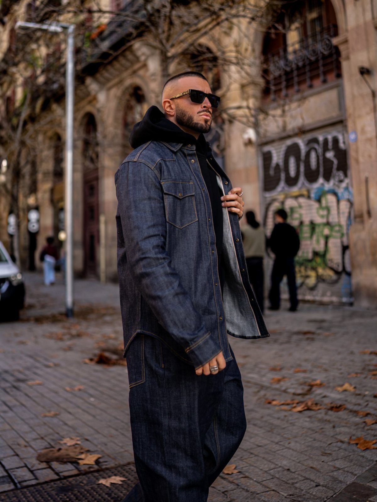 Man in dark denim jacket and matching trousers standing on a city street with autumn leaves.