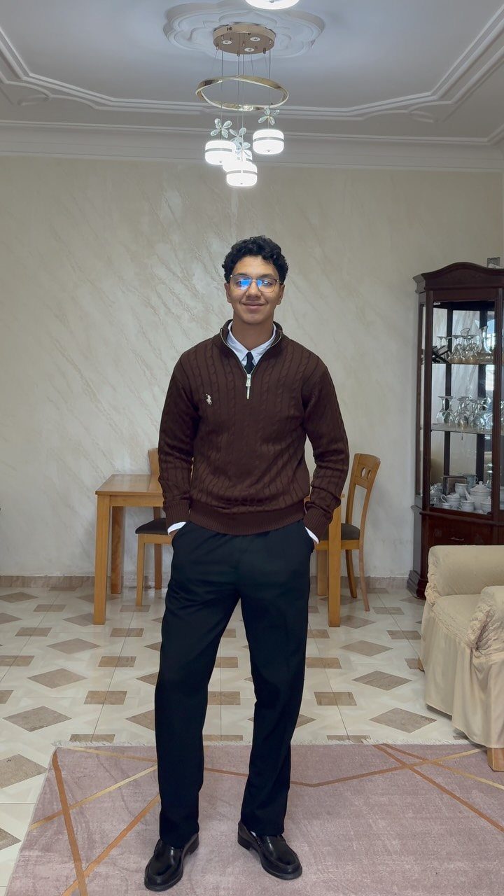A man wearing a brown quarter-zip sweater and black trousers standing in a living room.