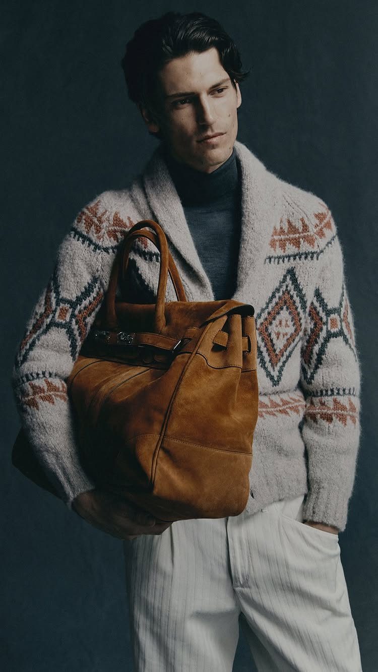 Man wearing a patterned knit cardigan and dark turtleneck while holding a large brown leather bag.