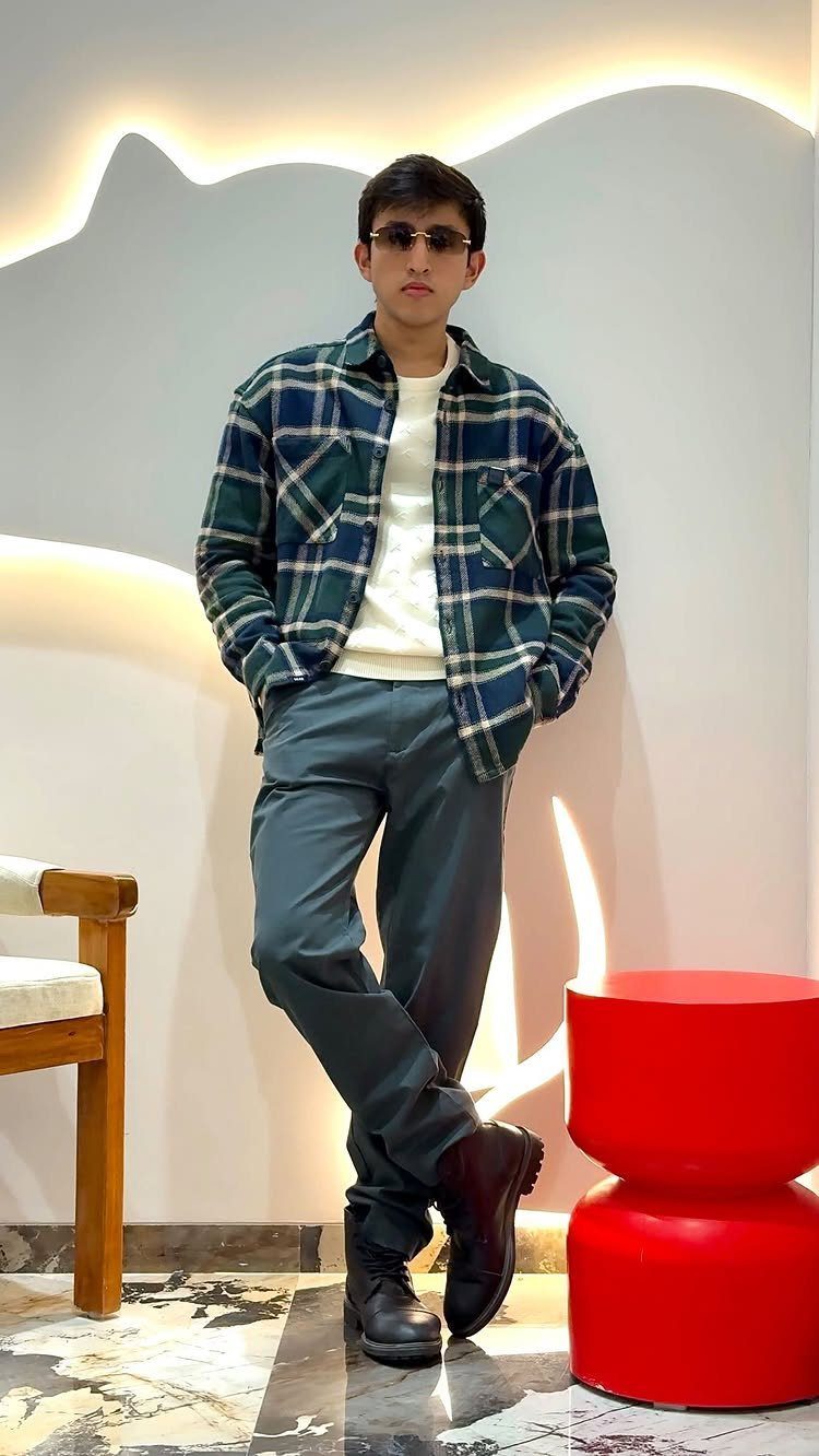 Man wearing a green plaid flannel shirt, white sweater, and dark trousers standing against a lit wall.